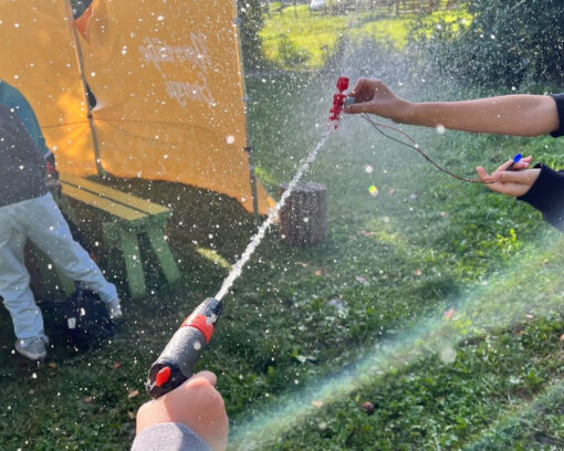 En närbild på två barnhänder som spritar vatten på varandra utomhus, grönt gräs i bakgrunden och solen slår igenom sprutet av vatten. Livligt.