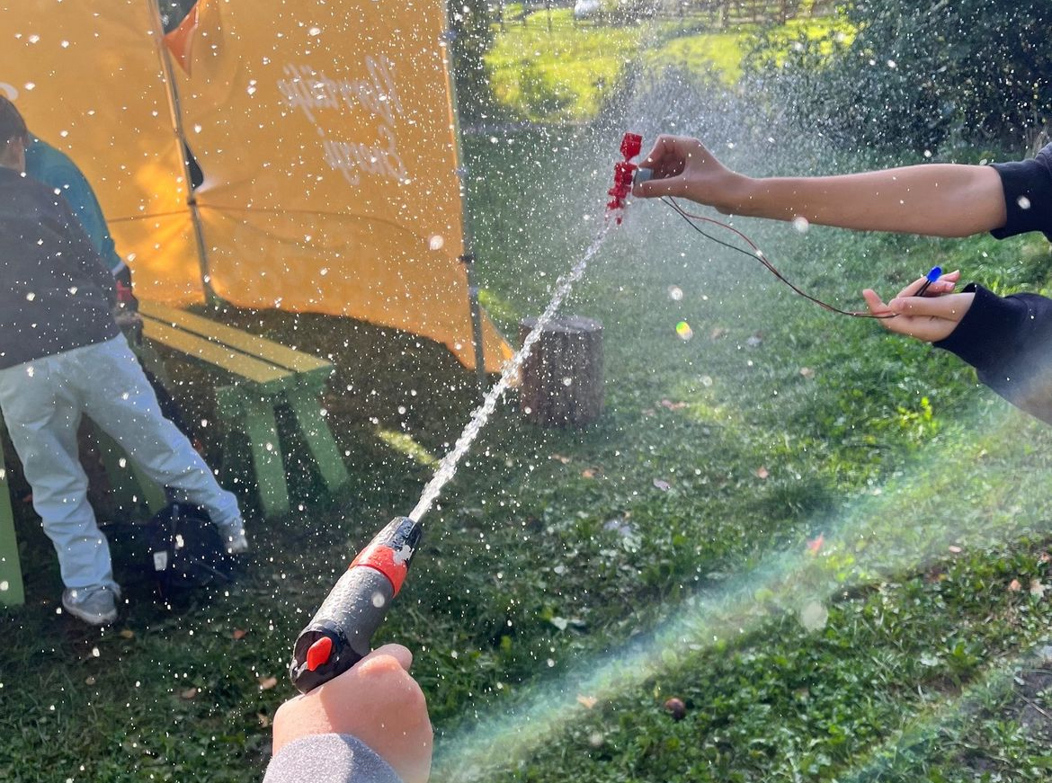 En närbild på två barnhänder som spritar vatten på varandra utomhus, grönt gräs i bakgrunden och solen slår igenom sprutet av vatten. Livligt.