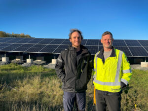 Frode och Tobias står med solelparken bakom sig, de ser in i kameran och ser nöjda ut. Utomhus med blå bakgrund och grönt gräs. 